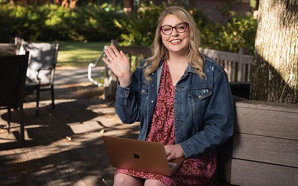 Admissions counselor Grace Tennyson smiles and waves while using a laptop and sitting on a bench.