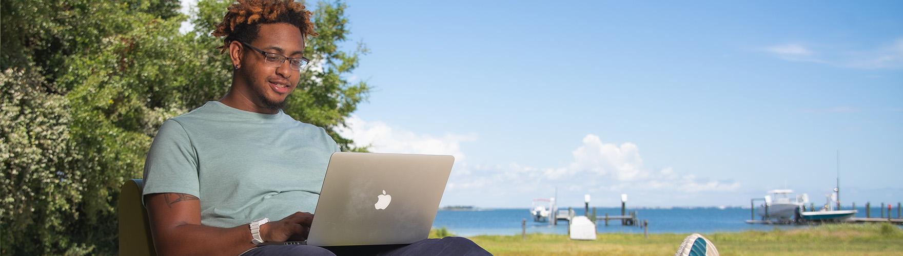 A student uses a laptop in a backyard by the water.