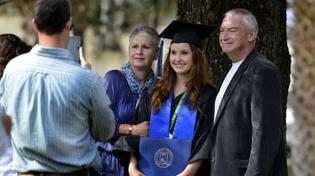 Female graduate is in her cap and gown holding her diploma and posing for a picture with her parents.